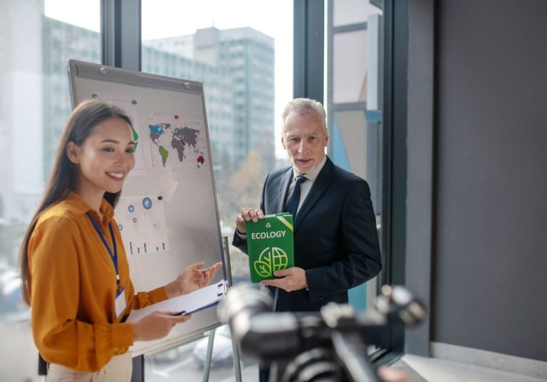 Mulher e homem conversam em frente a um quadro com mapas. O homem está segurando um livro de capa verde. Curso: Gestão Ambiental.
