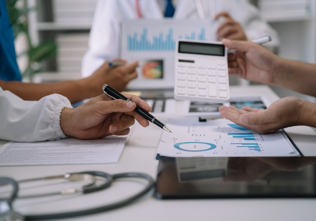 Profissionais analisam gráficos e documentos em mesa com estetoscópio e tablet. Curso: Gestão de Custos Hospitalares.