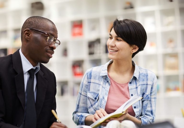 Homem e mulher conversam em uma biblioteca, a mulher segura um livro aberto. Curso: Habilidade de Escutar.