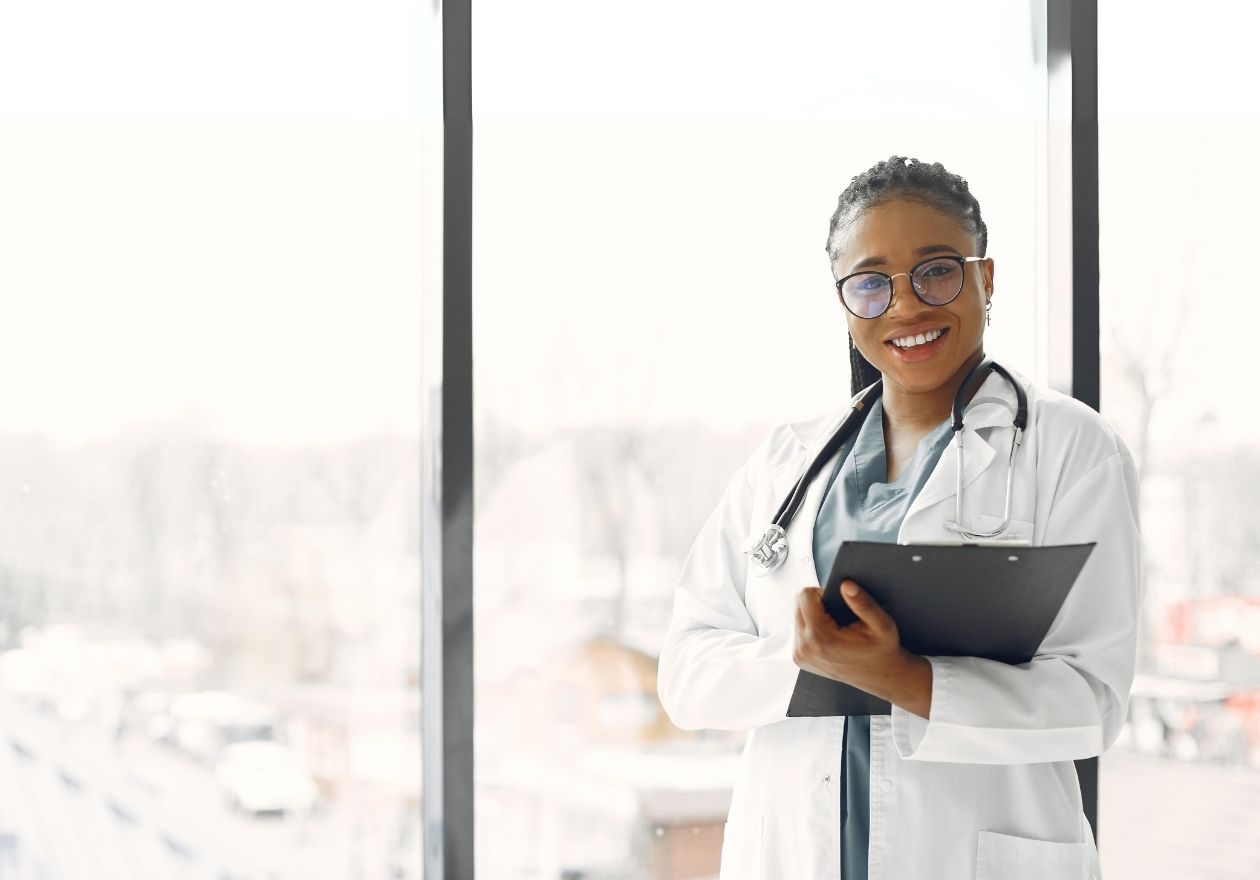 Uma profissional da saúde com jaleco branco segura um prontuário.Ela está sorrindo em um ambiente claro com janelas ao fundo. Curso: Interpretação dos Requisitos da RN 507/2022.