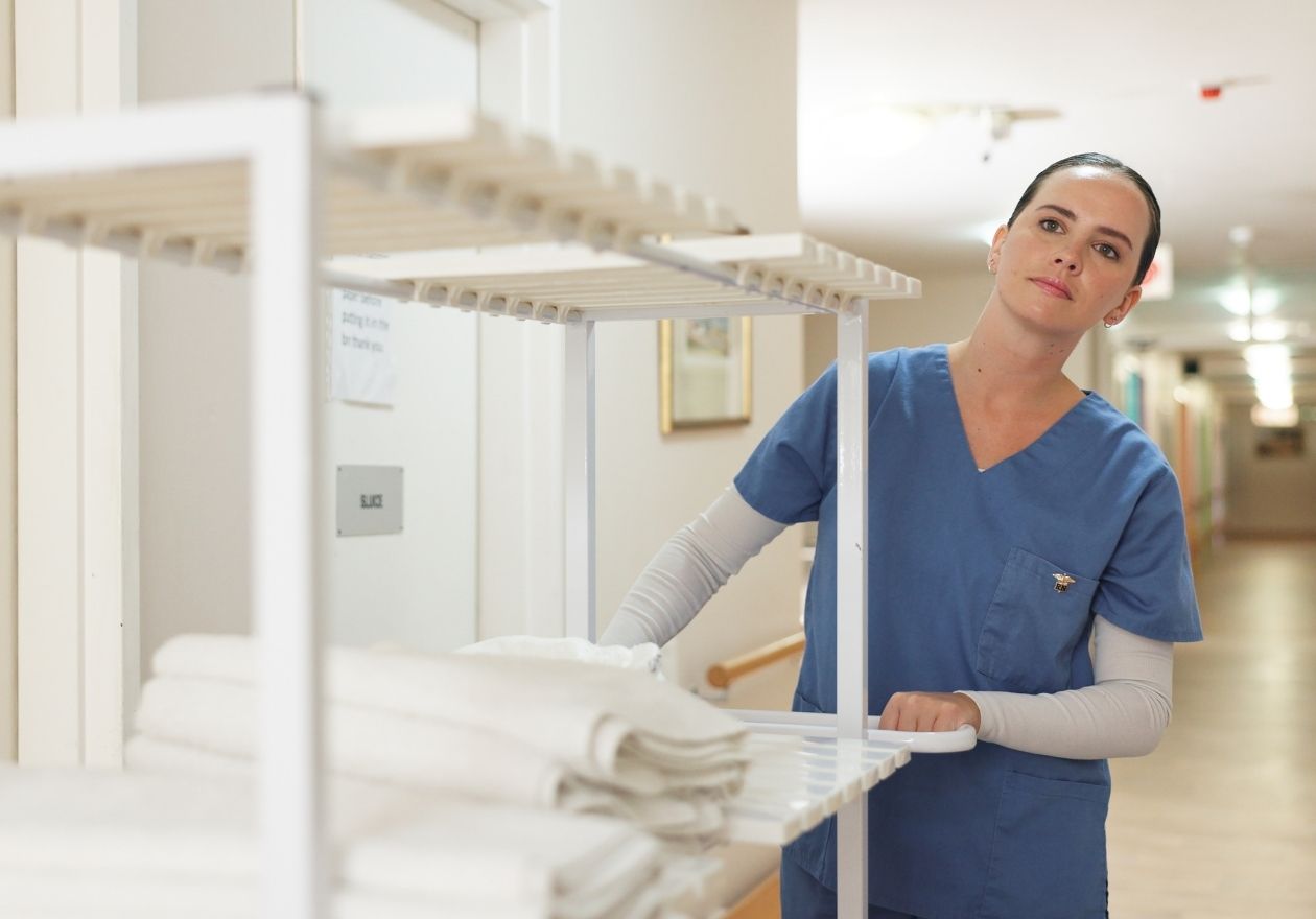 Mulher, profissional da saúde em uniforme azul organiza toalhas brancas em uma prateleira de hospital. Curso: Gestão de Lavanderias Hospitalares.
