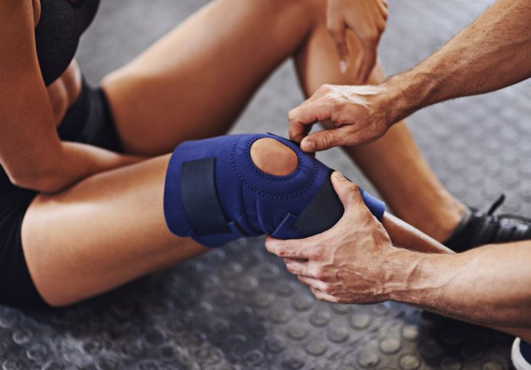 Profissional ajusta uma joelheira azul no joelho de uma mulher, em um ambiente de reabilitação ou treino. Curso: Lesões Meniscais.
