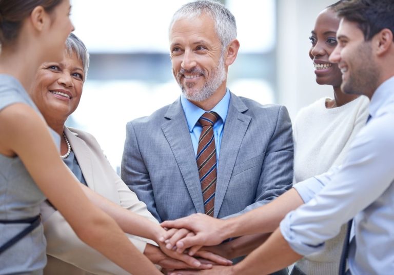Cinco pessoas unem as mãos em círculo, sorrindo, em um ambiente profissional. Curso: Liderança e Desenvolvimento de Equipes em Cooperativas.