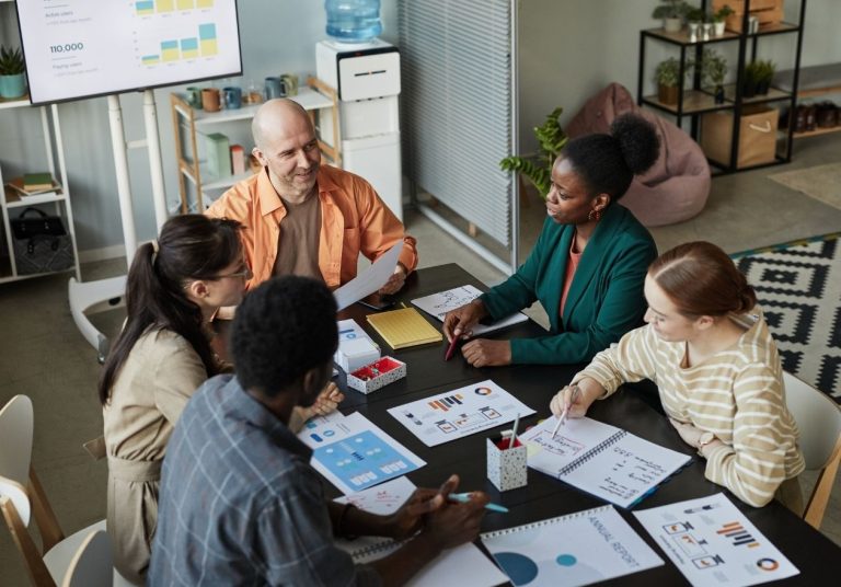 Cinco pessoas discutem ao redor de uma mesa com gráficos e documentos, em sala com quadro e plantas. Curso: Liderança e Gestão.