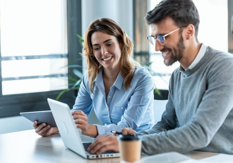 Duas pessoas sorridentes trabalham no laptop e tablet em um escritório com planta ao fundo. Curso: Mentoring