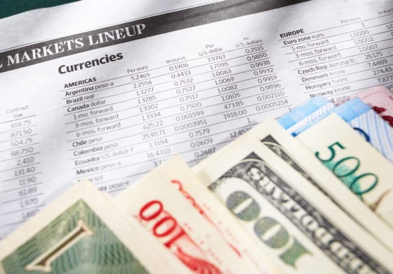 Notas de diferentes moedas e um jornal financeiro sobre uma mesa. Curso: Mercado de Câmbio - Abordagem Estratégica.