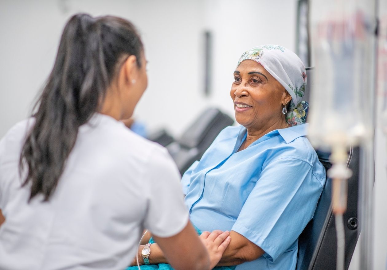 Uma profissional da saúde conversa com uma paciente idosa em um hospital. Curso: Navegação do Paciente Oncológico.