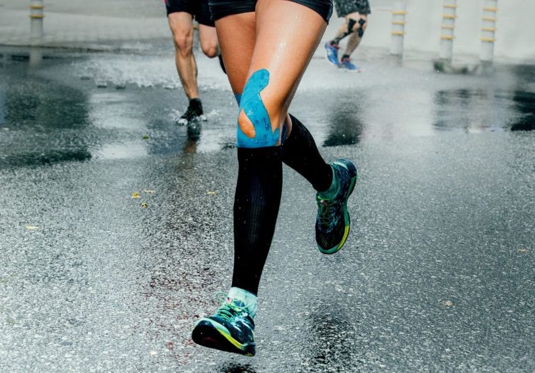 Uma corredora com joelheira azul corre em uma pista molhada, com outros corredores ao fundo. Curso: O Joelho no Esporte.