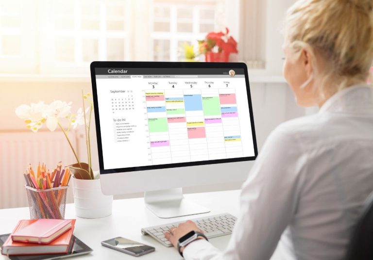 Uma mulher trabalha em um computador exibindo um calendário colorido, há flores e canetas ao lado. Curso: Organização Digital.