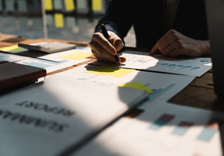 Mãos escrevem em papéis com gráficos e anotações, com post-its amarelos e um caderno sobre a mesa. Curso: Os 6 Sigmas.