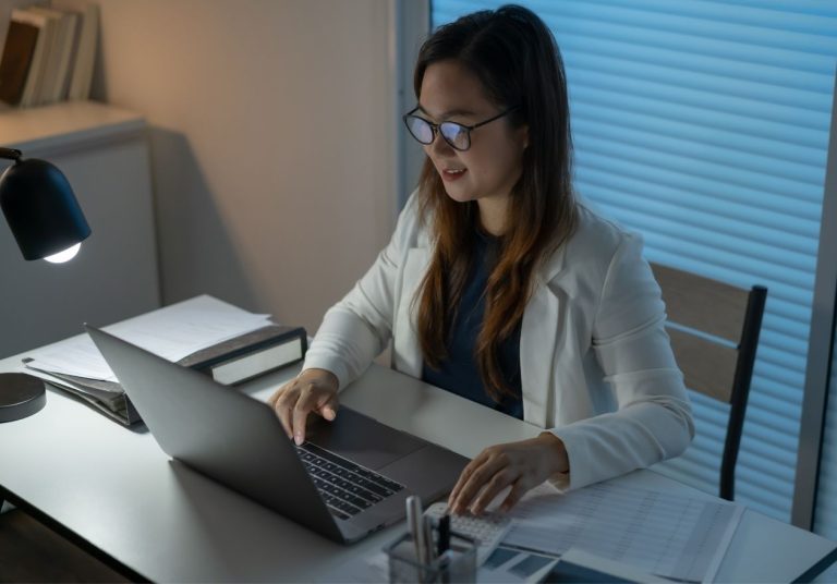 Mulher trabalha em um laptop em um escritório com papéis e uma luminária ao lado. Curso: Os 7 Pontos-Chave para a Produtividade.