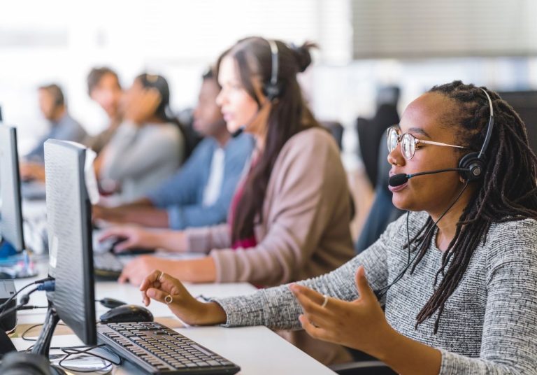 Funcionários com headsets atendem clientes em um escritório com vários computadores e colegas ao fundo. Curso: Ouvidoria - Princípios e Fundamentos da Função.