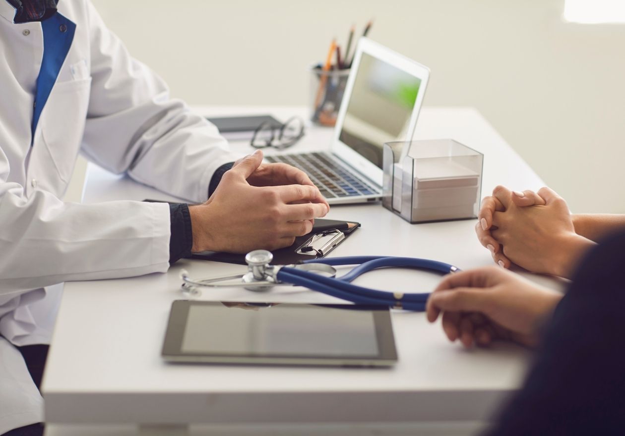 Um médico de jaleco conversa com um paciente, há estetoscópio, tablet e laptop na mesa. Curso: Programa de Capacitação em Governança Clínica.