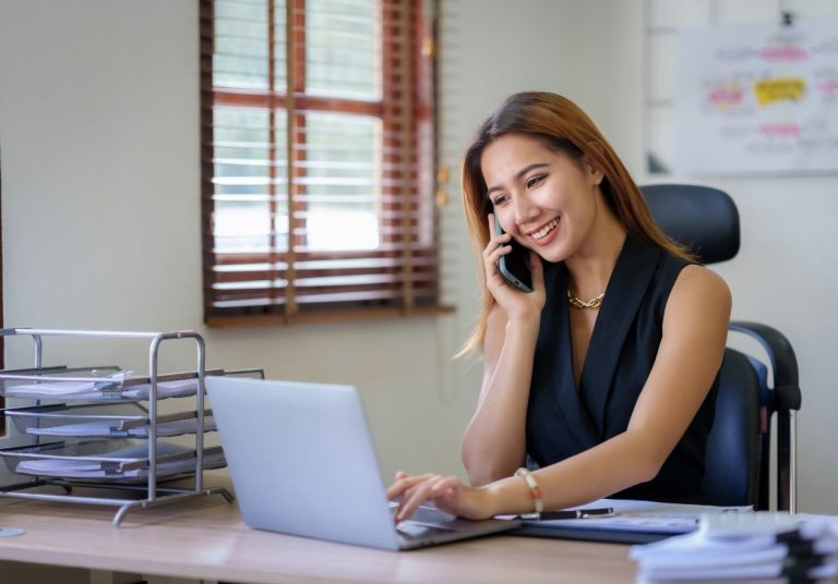Uma mulher sorridente fala ao telefone enquanto trabalha no laptop em um escritório. Curso: Programa Ouvidoria de Excelência.