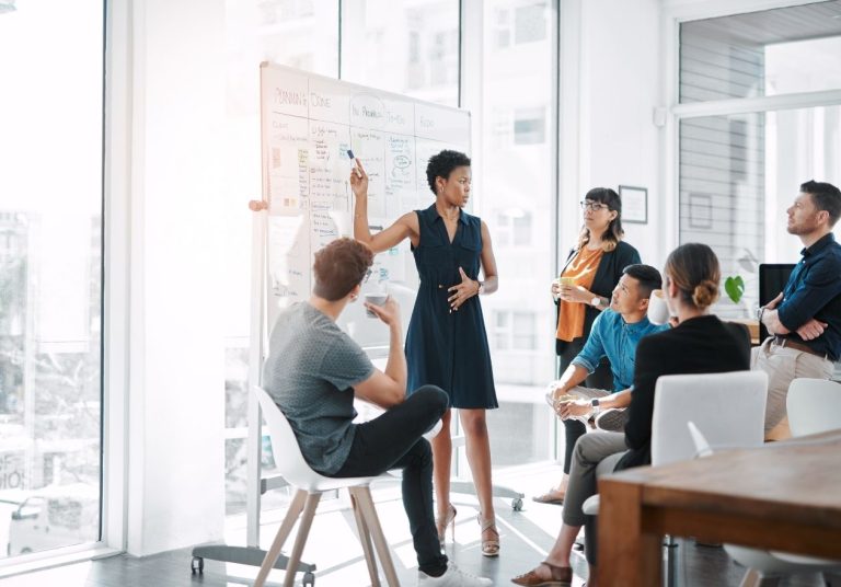 Uma mulher apresenta gráficos em um quadro para um grupo em sala de reunião. Curso: Protocolos da Implantação ao Gerenciamento.