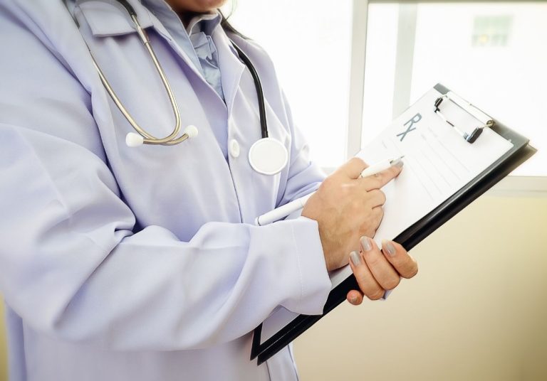 Um médico de jaleco segura uma prancheta e usa um estetoscópio no pescoço. Curso: Qualidade e Segurança.