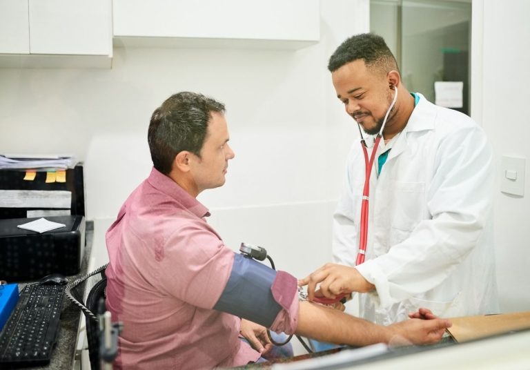 Médico em pé medindo pressão de paciente sentado, em consultório com estetoscópio e equipamentos. Curso: Qualificação dos Serviços AIS-APS.