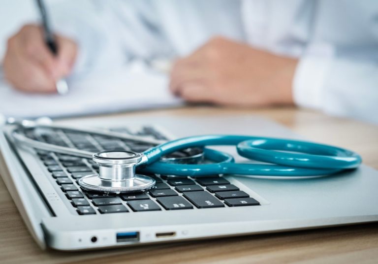 Estetoscópio azul sobre laptop, mãos ao fundo escrevendo em papel. Curso: Regulação dos Planos de Saúde - Obrigações das Operadoras.