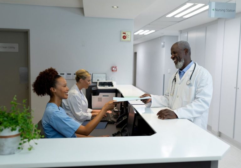 Médico e recepcionistas em balcão, com documentos e planta ao lado. Curso: Resultados e Melhorias.