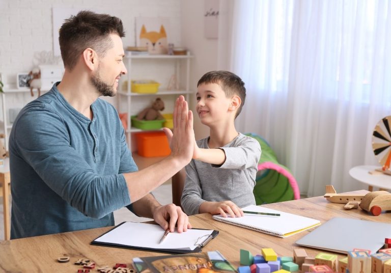 Adulto e criança dando "high five" em sala com brinquedos e materiais educativos. Curso: Sensibilização sobre TEA para Dirigentes do Sistema Unimed.