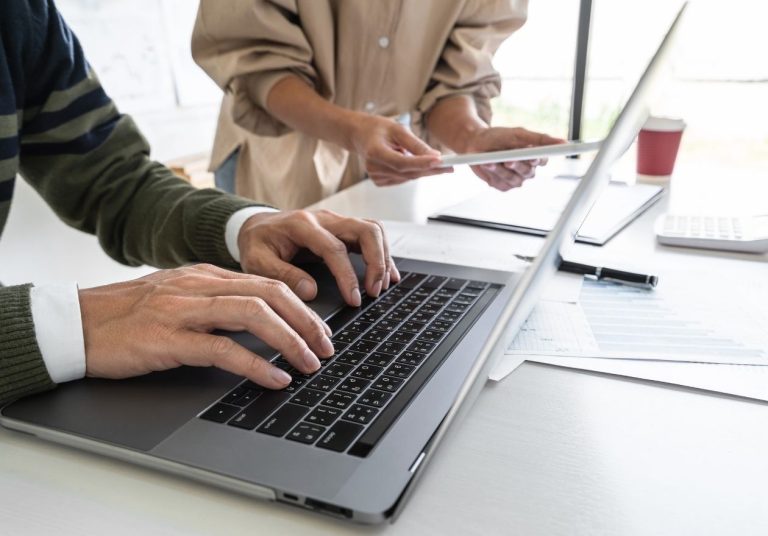 Pessoa digitando em um laptop, com outra ao fundo segurando papéis, em um ambiente de trabalho. Curso: Tecnologias Habilitadoras e Impacto dos Dados.