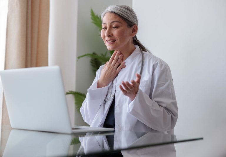 Médica em jaleco branco, gesticulando na frente de um laptop, em um ambiente claro com planta ao fundo. Curso: Telemedicina.