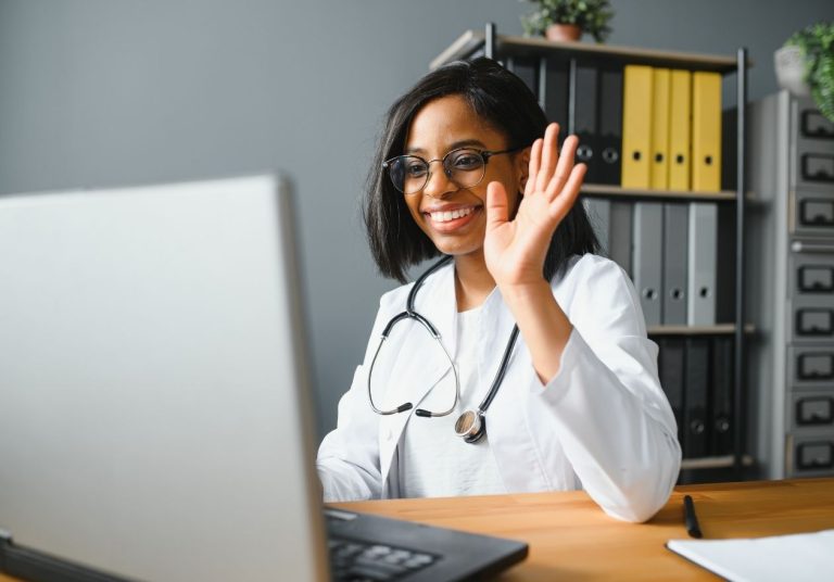Médica de jaleco, com estetoscópio, acenando para a câmera, usando laptop, com estante de arquivos ao fundo. Curso: Telemedicina em Saúde Mental.
