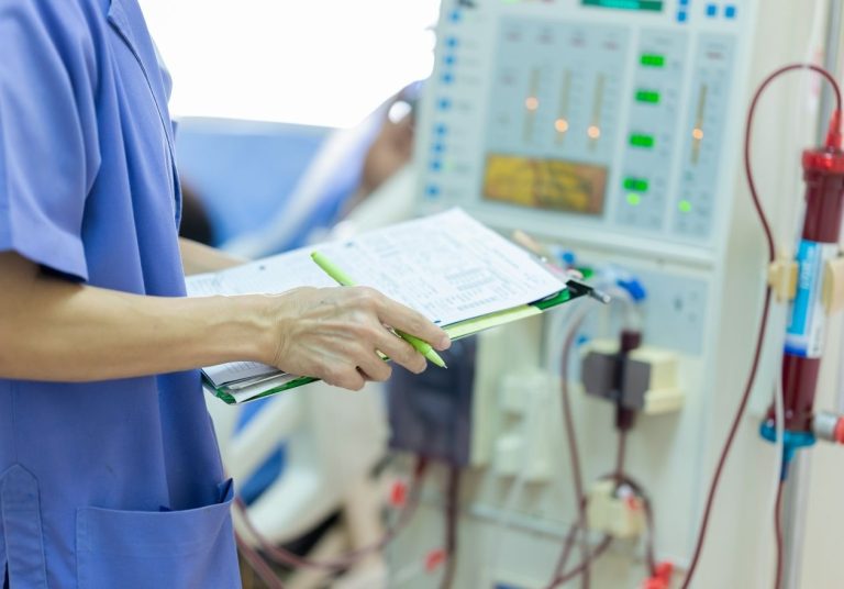 Profissional da saúde em uniforme azul, segurando prancheta, ao lado de equipamento de hemodiálise. Curso: Terapia Renal Substitutiva.
