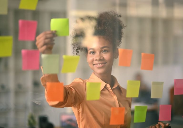 Mulher sorridente em frente a um vidro com post-its coloridos, organizando ideias. Curso: Traçar Metas e Objetivos Realistas.