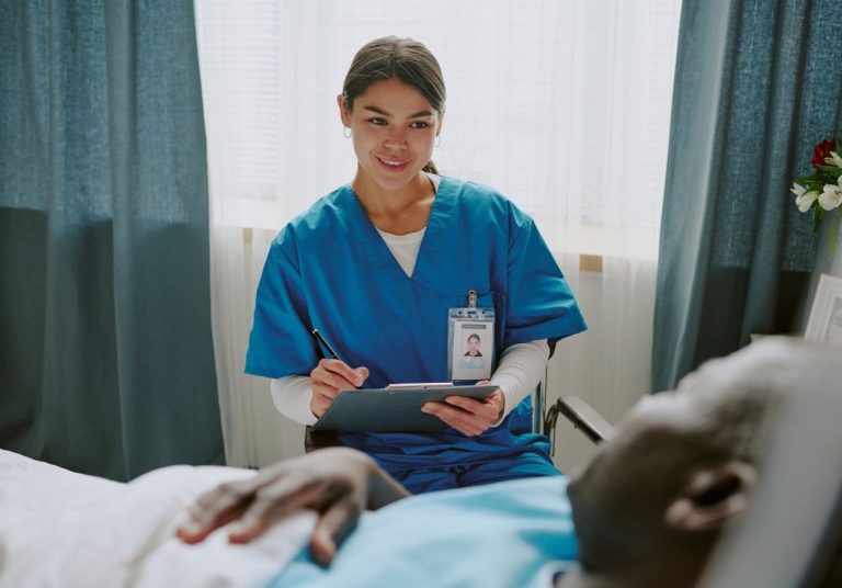 Enfermeira em uniforme azul, anotando algo em uma prancheta, ao lado de paciente deitado em cama hospitalar. Curso: Trajetória do Paciente.