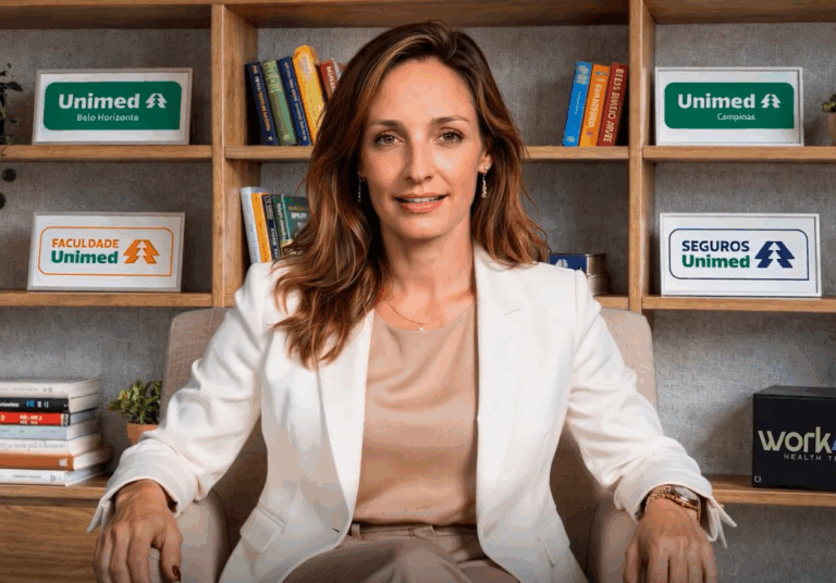 Avatar de IA da Dra Maria Ines desevolvido pelo projeto extensia. Reproduz uma mulher adulta branca e de cabelo loiro escuro, sentada de frente para uma câmera em um escritório e com paletó branco. Atrás tem uma estante com livros.